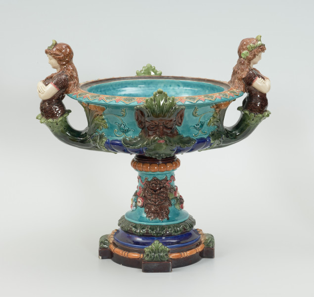 Waza - Ujęcie z tyłu. Waza w kształcie smukłej amfory. Brzusiec ceramiczny, rozszerzający się ku górze, w kolorze kremowym, malowany podszkliwnie (z uzupełnieniami naszkliwnymi) we wzory kwiatowe - bukiety barwnych kwiatów w typie aster w kolorach fioletowo-czerwonym, liliowym, żółtym i białym, z gałązkami i listkami oraz drobnymi kwiatkami przypominającymi bratki o barwie fioletowo-czerwonej i liliowej. Górna część wazy wraz z uchwytami oraz stopą i część podstawy wykonane jako odlewy z mosiądzu. Mosiężna szyja wąska z otokiem perełkowym w środku ozdobiona jest pionowym, rozszerzającym się ku dołowi i ku górze rytmicznym podziałem z ornamentem geometryczno-roślinnym. Na pogrubionym skraju szyi umieszczona jest płytka miseczka w formie kwiatu o wąskich płatkach, stanowiąca wylewkę wazonu. Poniżej dolnej części szyi oprawa wierzchołka brzuśca składająca się z otoku o sercowym wycięciu zwieńczonym profilowaną palmetką oraz dwóch wydłużonych, sięgających 1/3 wysokości brzuśca, pionowych wsporników uchwytów. Te ostatnie składają się z 2 części: zewnętrzna wychodząca ze wsporników opartych na brzuścu, wyniesiona jest po niewielkim skosie do połowy wysokości szyi i na końcu wywinięta do wnętrza wolutowo, a druga - krótsza, skierowana ku dołowi łączy łagodnym łukiem wolutę z górną częścią oprawy brzuśca, kończąc się wywiniętymi na zewnątrz listkami. Uchwyty żłobkowane, zdobione motywem liści. Wazon wsparty jest na mosiężnej podstawie, składającej się z wieńca oplecionego wstęgą, na którym spoczywa zwężająca się ku górze stopa o brzegach ozdobionych perełkowaniem i pierścieniem, wspierającym koszyczek, ujmujący dolną część ceramicznego brzuśca, w formie kwiatu o ostro zakończonych, wąskich płatkach. Całość spoczywa na podstawie z kwadratowej płytki czerwonego marmuru ze ściętymi narożnikami, ujętymi mosiężnymi wspornikami ramy wspierającej płytę od dołu i opartej w narożach na czterech lwich łapach.