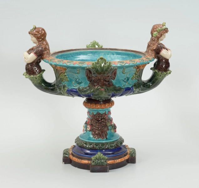 Waza - Ujęcie z tyłu. Waza w kształcie smukłej amfory. Brzusiec ceramiczny, rozszerzający się ku górze, w kolorze kremowym, malowany podszkliwnie (z uzupełnieniami naszkliwnymi) we wzory kwiatowe - bukiety barwnych kwiatów w typie aster w kolorach fioletowo-czerwonym, liliowym, żółtym i białym, z gałązkami i listkami oraz drobnymi kwiatkami przypominającymi bratki o barwie fioletowo-czerwonej i liliowej. Górna część wazy wraz z uchwytami oraz stopą i część podstawy wykonane jako odlewy z mosiądzu. Mosiężna szyja wąska z otokiem perełkowym w środku ozdobiona jest pionowym, rozszerzającym się ku dołowi i ku górze rytmicznym podziałem z ornamentem geometryczno-roślinnym. Na pogrubionym skraju szyi umieszczona jest płytka miseczka w formie kwiatu o wąskich płatkach, stanowiąca wylewkę wazonu. Poniżej dolnej części szyi oprawa wierzchołka brzuśca składająca się z otoku o sercowym wycięciu zwieńczonym profilowaną palmetką oraz dwóch wydłużonych, sięgających 1/3 wysokości brzuśca, pionowych wsporników uchwytów. Te ostatnie składają się z 2 części: zewnętrzna wychodząca ze wsporników opartych na brzuścu, wyniesiona jest po niewielkim skosie do połowy wysokości szyi i na końcu wywinięta do wnętrza wolutowo, a druga - krótsza, skierowana ku dołowi łączy łagodnym łukiem wolutę z górną częścią oprawy brzuśca, kończąc się wywiniętymi na zewnątrz listkami. Uchwyty żłobkowane, zdobione motywem liści. Wazon wsparty jest na mosiężnej podstawie, składającej się z wieńca oplecionego wstęgą, na którym spoczywa zwężająca się ku górze stopa o brzegach ozdobionych perełkowaniem i pierścieniem, wspierającym koszyczek, ujmujący dolną część ceramicznego brzuśca, w formie kwiatu o ostro zakończonych, wąskich płatkach. Całość spoczywa na podstawie z kwadratowej płytki czerwonego marmuru ze ściętymi narożnikami, ujętymi mosiężnymi wspornikami ramy wspierającej płytę od dołu i opartej w narożach na czterech lwich łapach.
