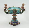 Waza - Ujęcie z tyłu. Waza w kształcie smukłej amfory. Brzusiec ceramiczny, rozszerzający się ku górze, w kolorze kremowym, malowany podszkliwnie (z uzupełnieniami naszkliwnymi) we wzory kwiatowe - bukiety barwnych kwiatów w typie aster w kolorach fioletowo-czerwonym, liliowym, żółtym i białym, z gałązkami i listkami oraz drobnymi kwiatkami przypominającymi bratki o barwie fioletowo-czerwonej i liliowej. Górna część wazy wraz z uchwytami oraz stopą i część podstawy wykonane jako odlewy z mosiądzu. Mosiężna szyja wąska z otokiem perełkowym w środku ozdobiona jest pionowym, rozszerzającym się ku dołowi i ku górze rytmicznym podziałem z ornamentem geometryczno-roślinnym. Na pogrubionym skraju szyi umieszczona jest płytka miseczka w formie kwiatu o wąskich płatkach, stanowiąca wylewkę wazonu. Poniżej dolnej części szyi oprawa wierzchołka brzuśca składająca się z otoku o sercowym wycięciu zwieńczonym profilowaną palmetką oraz dwóch wydłużonych, sięgających 1/3 wysokości brzuśca, pionowych wsporników uchwytów. Te ostatnie składają się z 2 części: zewnętrzna wychodząca ze wsporników opartych na brzuścu, wyniesiona jest po niewielkim skosie do połowy wysokości szyi i na końcu wywinięta do wnętrza wolutowo, a druga - krótsza, skierowana ku dołowi łączy łagodnym łukiem wolutę z górną częścią oprawy brzuśca, kończąc się wywiniętymi na zewnątrz listkami. Uchwyty żłobkowane, zdobione motywem liści. Wazon wsparty jest na mosiężnej podstawie, składającej się z wieńca oplecionego wstęgą, na którym spoczywa zwężająca się ku górze stopa o brzegach ozdobionych perełkowaniem i pierścieniem, wspierającym koszyczek, ujmujący dolną część ceramicznego brzuśca, w formie kwiatu o ostro zakończonych, wąskich płatkach. Całość spoczywa na podstawie z kwadratowej płytki czerwonego marmuru ze ściętymi narożnikami, ujętymi mosiężnymi wspornikami ramy wspierającej płytę od dołu i opartej w narożach na czterech lwich łapach.