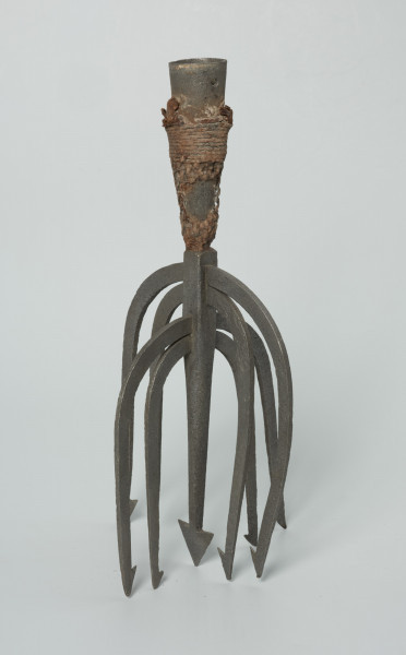 Ość dziewięciozębna kandelabrowa - Ujęcie z przodu skosem w lewą stronę. Ość dziewięciozębna, wykonana z kutego żelaza. Oś narzędzia stanowi ząb z trójkątnym grotem, zakończony oplecioną konopnym sznurkiem stożkowatą tuleją. W górnej części tulei dwa niewielkie, umieszczone naprzeciwlegle względem siebie otwory.  Ząb o zwężających się ku dołowi ściankach. W górnej i dolnej części mający przekrój prostokątny, w środkowej zbliżony do okrągłego. Blisko jego nasady znajdują się, na prostopadłych względem siebie ściankach, dwa przechodzące na wylot prostokątne otwory, w których mocowane są po dwie pary łukowato wygiętych ku dołowi ramion (pozostałych zębów) zakończonych pojedynczymi zadziorami (jednego zadziora brak). Ułożenie ramion nadaje narzędziu formę kandelabrową.