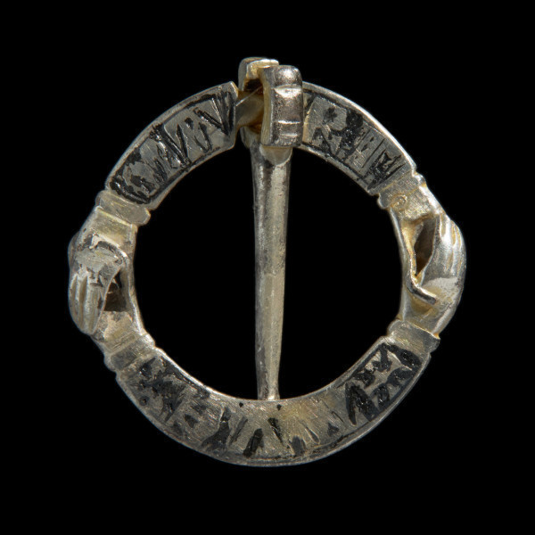 Pozłacana zapinka ze splecionymi dłońmi - Ujęcie z przodu w pionie. Pozłacana zapinka, wykonana z dwóch owalnych taśm, w końcach uformowanych w dłonie. Powierzchnie zdobione motywem krzyży i napisami  AVE MARI. Kolec wąsk, długi.