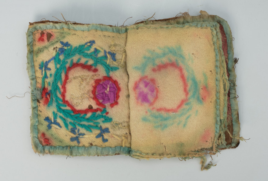 książeczka na igły z haftowanymi okładkami i kartkami z wełny - Ujęcie środka obiektu, pierwsza strona; Przedmiot w kształcie otwartej książeczki, w której karty wykonane są z miękkiej, wełnianej tkaniny w kolorze naturalnym. Na lewej karcie płaski, prosty haft w kolorze niebieskim, fioletowym i czerwonym. Na drugiej karcie brak haftu. Obie karty z obszytymi brzegami jasnoniebieską włóczką.