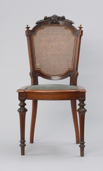 krzesło - Ujęcie z przodu. Krzesło z płasko wyściełanym siedziskiem i oparciem z plecionki z rafii. Przednie nogi toczone, tralkowe, w części środkowej wydzielonej wałkami - kanelowane. Tylne nogi o przekroju okrągłym, lekko wygięte, przechodzące w prostokątne słupki zaplecka. Siedzenie w kształcie podkowiastym, z przodu lekko zaokrąglone na zewnątrz, w środku wyściełane - tkanina mocowana pinezkami tapicerskimi. Na styku przednich nóg i ramy siedziska ozdobna kostka z płycinami na obu ściankach zewnętrznych. Rama zaplecka w formie medalionu, wypełnionego plecionką z rafii. Słupki zaplecka dekorowane wgłębnym reliefem w formie płycin i spirali, a łuk zwieńczenia zdobi dekoracja o formach rollwerkowo-roślinnych. Ponad kostkami wieńczącymi boczne ramy zaplecka toczone gałki. Siedzisko wyściełane tkaniną w kolorze zielonym (turkusowym?), z brzegami wykończonymi pasmanterią.