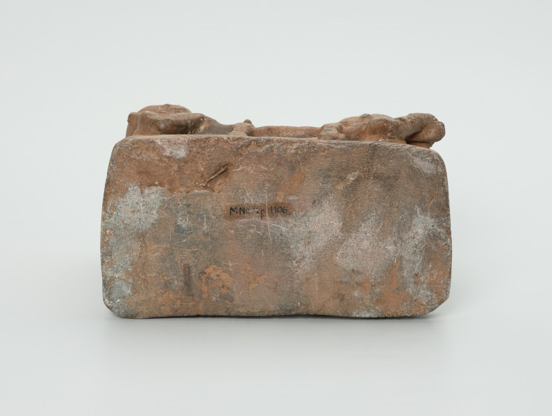 figura kultu przodków - Ujęcie spodu figury. Rzeźbiona w brązowo-szarym kamieniu scena przedstawiająca naczelnika niesionego w lektyce przez czterech niewolników. Części twarzowe wszystkich postaci mają cechy negroidalne: wydatne, lekko rozchylone usta oraz szeroki u nasady nos. Oczy przymknięte, zamyślone. Uszy wyrzeźbiono na wysokości oczu niewolnikom, z tym wyjątkiem, że dwóm tylnym jedynie po jednym od wewnętrznej strony. Naczelnik nie ma uszu. Na twarzach postaci widoczne bruzdy na czole. Naczelnikowi zaznaczono również po dwa nacięcia na policzkach. Ręce naczelnika zgięte w łokciach, a dłonie oparte na drągach lektyki. Jego tył głowy i plecy przylegają do oparcia lektyki, natomiast zgięte w kolanach nogi ułożono jedna na drugą. Niewolników przedstawiono w pozycji stojącej. Ich powieki ozdobiono ukośnymi nacięciami. Ręce niewolników zgięte w łokciach, w dłoniach trzymają drąg od lektyki, który oparto na jednym z ich ramion. Ich nogi złączono, zaznaczono stopy. Rzeźbę umieszczono na prostokątnej podstawie, którą ozdobiono ukośnymi nacięciami.