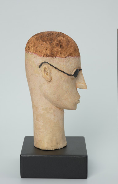 lalka teatralna: Tubabu Cè Kun - Ujęcie z prawego boku. Drewniane popiersie mężczyzny.  Głowa owalna, wydłużona, zaznaczona linia włosów nad czołem. Włosy pomalowane na kolor jasno-brązowy/blond. Nos długi, wąski, usta wypukle, pomalowane na kolor jasno-różowy. Zamiast oczu czarne okulary, okrągłe, z cienkimi nausznicami. Spod okularów wystające powieki dolne i górne. Szyja długa, wąska, rozszerzona u dołu, pomalowana na kolor jasno-różowy.