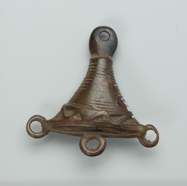 ozdoba ciała , biżuteria - Ujęcie z przodu. Mały, trójkątny wisiorek, wykonany z brązu. Całość ozdobiona wzorami geometrycznymi. W dolnej części udekorowany trzema małymi kółeczkami, w górnej zaś części znajduje się mały i okrągły otwór.