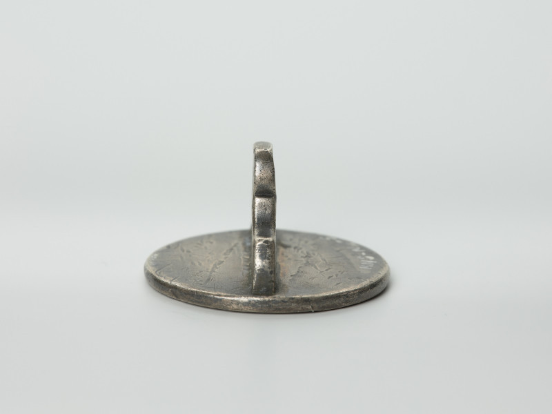 tłok pieczętny - Ujęcie z boku z lewej strony pieczęcią w dół. Okrągły tłok pieczętny ze srebra z uchwytem w formie profilowanej płytki oraz wizerunkiem łba gryfa i gotyckim napisem w polu pieczęci.