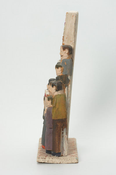 rzeźba - Ujęcie prawej strony. Rzeźba wykonana w jednym kawałku drewna lipowego . Opracowana tylko od strony frontalnej. Wolnostojąca na prostokątnym postumencie jest , bryłą zwartą, słabo rozczłonkowaną, statyczną. Postacie umieszczone wertykalnie: w trzech rzędach sami mężczyźni ubrani w garnitury, trzymający w rękach biało - czerwone i czerwoną chorągiewki. Powyżej kobiety ( widoczne w zasadzie same głowy) trzymają transparent z napisem: 