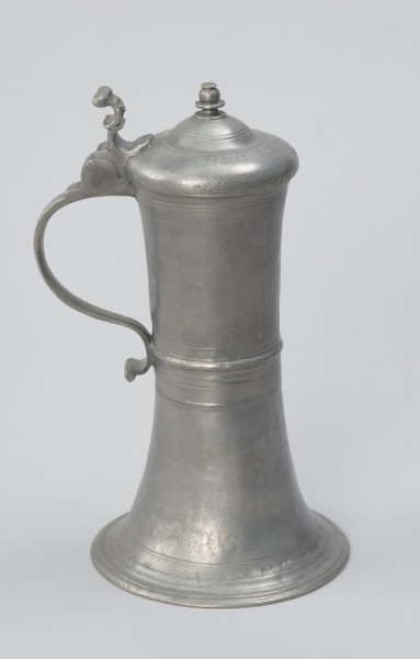 Cynowy dzban o kształcie cylindrycznym - Ujęcie z prawej strony; Wysmukły dzban o kształcie cylindrycznym z lekko wklęsłym brzuścem, z pierścieniowym zgrubieniem w centrum. Rozszerzająca się ku dołowi podstawa przechodzi w profilowaną stopę. Ucho taśmowe, silnie esowato wygięte, zakończone wałkiem. W górnej części ucha zawias podtrzymujący pokrywę z uchwytem wygiętym esowato. Pokrywa kopulasta, dwustopniowa z profilowaną, toczoną gałką do podnoszenia na szczycie. Na brzuścu u podstawy, pośrodku i przy górnej krawędzi, a także na pokrywie delikatne profilowane i ryte pasy.