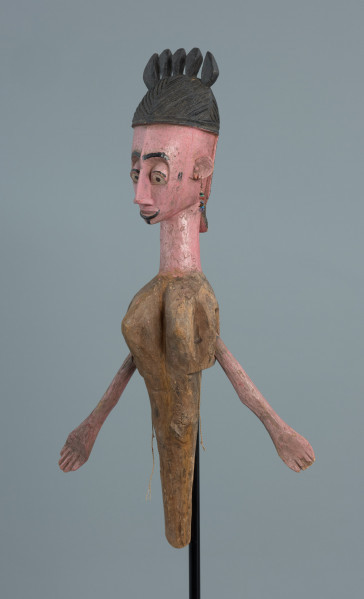 lalka teatralna: Yayoroba - Ujęcie z przodu skosem w lewą stronę. Drewniana lalka przedstawiająca postać kobiecą. Różowa karnacja, mocno zaznaczone brwi, powieki górne, białe oczy z czarnymi źrenicami. Czarne, wysoko upięte włosy z pięcioma wypustkami. Odstające uszy, długi nos i wąskie usta. Zaznaczona cienka czarna broda. Z uszu zwisają długie kolczyki z kolorowych koralików. Szyja jest  nieproporcjonalnie długa. Lalka ma na sobie brązową sukienkę.