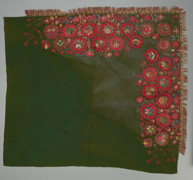 zielona chusta z, haftem z motywami kwiatowymi, cekinami i frędzlami - Ujęcie z przodu; Chusta kwadratowa, z wełnianej, ciemnozielonej tkaniny, zdobiona haftem płaskim z motywem wielopłatkowych kwiatków w kolorze czerwonym, ułożonych w trzech rzędach, jeden nad drugim, w rzędzie dolnym kwiatki mniejsze. Środki kwiatów o kolorach niebieskim, żółtym i zielonym układają się w różne wzory florystyczne. Każdy z kwiatów obwiedziony jest złotym sznureczkiem. Pomiędzy większymi dodatkowo wyhaftowane są drobniejsze kwiatki. całość zdobiona także cekinami. Pomiędzy kwiatami w rogu napis: 