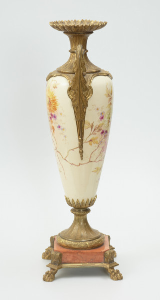 Waza - Ujęcie z lewej strony Waza dekoracyjna o bogatej, barwnej dekoracji reliefowej, w kształcie kielicha ze spłaszczoną czarą z dwoma uchwytami i przykrywą. Podstawa okrągła, profilowana z czterema prostokątnymi poszerzeniami z listkiem akantu. Trzon na styku z podstawą ozdobiony wałkiem z liści akantu, w części środkowej wypełniony reliefem z dwiema brązowymi maskami i girlandami drobnych, barwnych kwiatów. W górnej części trzonu perełkowy otok z przewężeniem na styku z czarą. Czara z szerokim brzegiem ozdobionym dekoracją geometryczną i zwężającym się do dołu brzuścem dekorowanym dwiema wypełniającymi całą wysokość czary brązowymi maskami, których zielone grzywy w formie palmety opierają się na górnej krawędzi brzegu, a brody na spodzie czary ozdobionym granatowymi 