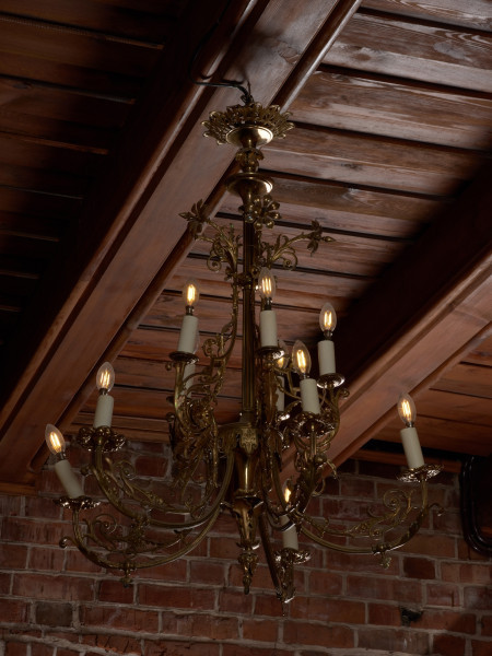 żyrandol - Ujęcie z prawej strony. Żyrandol 12-ramienny, mosiężny z kryształowymi soplami.Trzon w górnej części o przekroju kwadratowym, ozdobiony wypukłą dekoracją o formach geometrycznych i roślinnych, w dolnej części tralkowy z zakończeniem w kształcie szyszki. Ramiona w formie gałązek wygiętych półkoliście, połączonych przy trzonie w 4 wiązki po 3 elementy: dwa dłuższe po bokach i krótsze w środku. Każda trójka przechodzi przy trzonie w jedną spiralnie zwiniętą gałązkę akantu. Środkowe części ramion ozdobione są analogicznymi motywami: łodyżką z listkami rozszczepiającą się na dwa spiralnie skręcone pędy. Profitki ażurowe, zbudowane z ułożonych w rozetkę listków akandu. Przy górnej części trzonu, w połowie wysokości motyw dekoracyjny złożony z dwóch wygiętych, a na końcach spiralnie skręconych gałązek akantu. Górny talerz maskujący pod sufitem zawieszkę - płaski w formie kwiatu akantu. Ramiona i trzon połączone luźno zwieszonymi łańcuszkami, umieszczonymi także przy talerzu podsufitowym i ozdobach górnej części trzonu. Przy profitkach szklane wisiorki: 16 sztuk szlifowanych w kształcie sopelków - podłużne o trójkątnym przekroju z ostrosłupowym zakończeniem oraz 5 sztuk w kształcie pękatych łezek. Świece tulejowe z tworzywa sztucznego, zaopatrzone we wkręty do żarówek świecowych. Wewnętrzna instalacja elektryczna.