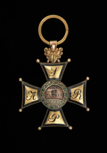 Order Virtuti Militari III klasy - Ujęcie pełnego awersu. Na awersie ramion napis. W okrągłej tarczy w centrum krzyża biało emaliowany orzeł w koronie z berłem i jabłkiem w łapach, otoczony wieńcem laurowym z zielono emaliowanymi liśćmi.