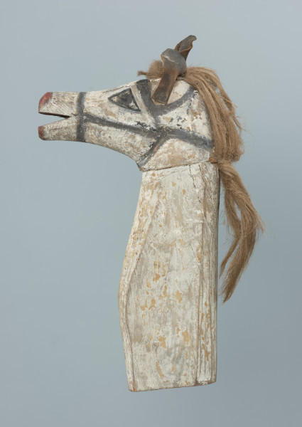 Głowa konia - rekwizyt obrzędowy - Ujęcie z prawej strony. Głowa konia – rekwizyt obrzędowy. Wyrób własny, jednostkowy. Głowa wykonana techniką obróbki stolarskiej z dwóch kawałków drewna łączonych na czop. Przedstawiona realistycznie. Pysk długi, otwarty. Jego środek oraz nozdrza pomalowane na czerwono (obecnie jedynie pozostałości farby). Z przodu żuchwy szereg bardzo słabo widocznych namalowanych białą farbą prostych kresek (zęby?).  Czarną farbą namalowane oczy i uzda. Po bokach głowy, za pomocą gwoździ, mocowane wycięte ze skóry uszy. Pomiędzy nimi  oraz poniżej, na karku, przybite za pomocą małych gwoździków cienkie skórzane paski, za pomocą których mocowana jest grzywa z jasnego włosia (końskiego?). Szyja długa, prosta, w jej spodzie znajduje się otwór na kij. Całość pomalowana na biało.