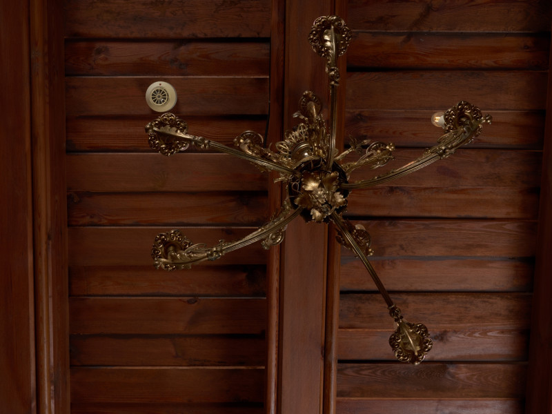 żyrandol - Ujęcie od dołu. Żyrandol 12-ramienny, mosiężny z kryształowymi soplami.Trzon w górnej części o przekroju kwadratowym, ozdobiony wypukłą dekoracją o formach geometrycznych i roślinnych, w dolnej części tralkowy z zakończeniem w kształcie szyszki. Ramiona w formie gałązek wygiętych półkoliście, połączonych przy trzonie w 4 wiązki po 3 elementy: dwa dłuższe po bokach i krótsze w środku. Każda trójka przechodzi przy trzonie w jedną spiralnie zwiniętą gałązkę akantu. Środkowe części ramion ozdobione są analogicznymi motywami: łodyżką z listkami rozszczepiającą się na dwa spiralnie skręcone pędy. Profitki ażurowe, zbudowane z ułożonych w rozetkę listków akandu. Przy górnej części trzonu, w połowie wysokości motyw dekoracyjny złożony z dwóch wygiętych, a na końcach spiralnie skręconych gałązek akantu. Górny talerz maskujący pod sufitem zawieszkę - płaski w formie kwiatu akantu. Ramiona i trzon połączone luźno zwieszonymi łańcuszkami, umieszczonymi także przy talerzu podsufitowym i ozdobach górnej części trzonu. Przy profitkach szklane wisiorki: 16 sztuk szlifowanych w kształcie sopelków - podłużne o trójkątnym przekroju z ostrosłupowym zakończeniem oraz 5 sztuk w kształcie pękatych łezek. Świece tulejowe z tworzywa sztucznego, zaopatrzone we wkręty do żarówek świecowych. Wewnętrzna instalacja elektryczna.