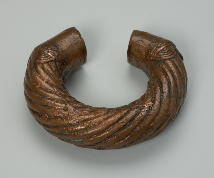 biżuteria, bransoleta - Ujęcie z tyłu z dołu. Otwarta, miedziana, owalna bransoleta ozdobiona grubymi, zaokrąglonymi, biegnącymi dookolnie prążkami; cienkimi, ukośnymi nacięciami oraz półkolami przypominającymi wachlarze wyłaniające się z motywu plecionki.