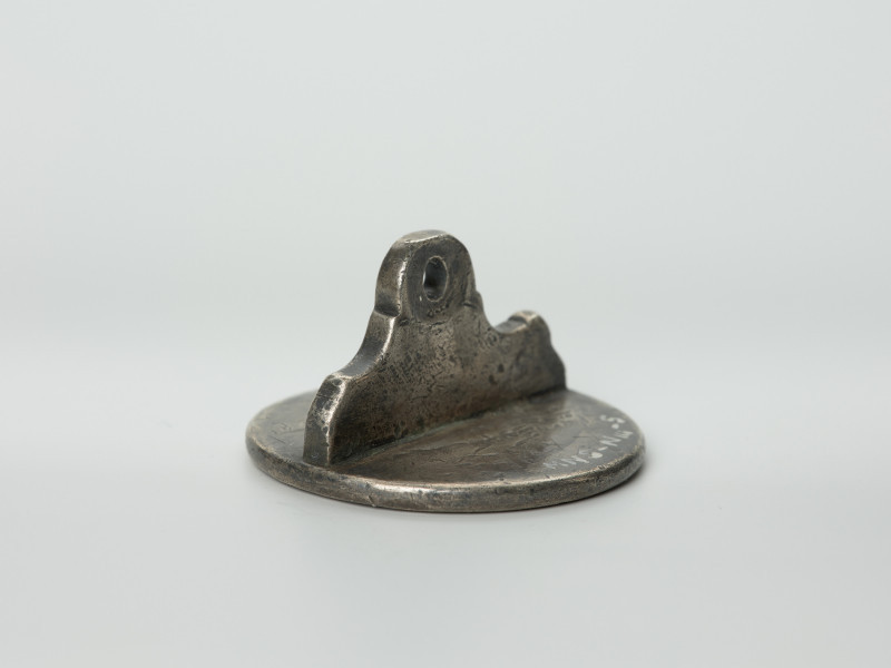 tłok pieczętny - Ujęcie z boku pieczęcia w dół. Okrągły tłok pieczętny ze srebra z uchwytem w formie profilowanej płytki oraz wizerunkiem łba gryfa i gotyckim napisem w polu pieczęci.