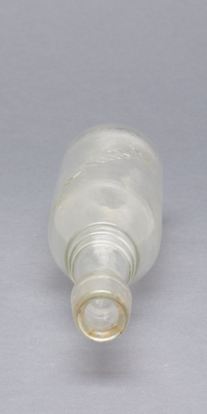 szklana butelka z wyodrębnioną szyjką z trzema pierścieniami - Ujęcie z góry; Szklana butelka piwówka z grubego szkła butelkowego półbiałego przezroczystego. Szyjka wyodrębniona trzema wypukłymi, wąskimi pierścieniami, umieszczonymi w dolnej części szyjki. U dołu pogrubionego wylewu pogrubienia na dwa otwory do zamknięcia pałąkowego. Dno zagłębione płaskie. Zamknięcie pałąkowe. Ślad szwu bocznego.