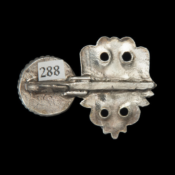 Pozłacana haftka z orłem dwugłowym - Ujęcie z tyłu. Pozłacana ozdoba z prostym orłem dwugłowym. Zaznaczone skrzydła i ogon. Po lewej stronie przyklejony okrągły, płaski element. Powierzchnia płaska.