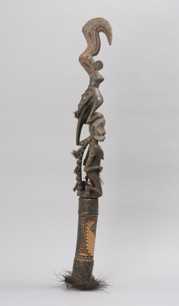 flet obrzędowy - Ujęcie z boku z prawej; Podłużny, bogato zdobiony flet wykonany z odcinka bambusa otwartego po stronie służącej do zadęcia i zamkniętego naturalnym kolankiem po stronie przeciwnej, gdzie znajduje się drewniana figura kobiety-przodka w pozycji klęczącej z ptakiem na głowie.