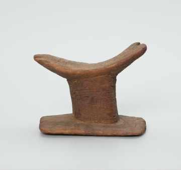 obrządek grzebalny, pochówek, podgłówek, rzeźba - Ujęcie z przodu. Rzeźbiony, drewniany podgłówek pokryty warstwą brunatnej ziemi. Część górna oparta na walcowatym dźwigarze. W jego dolnej części prostokątna podstawa. Widoczne liczne spękania oraz półokrągły ubytek.