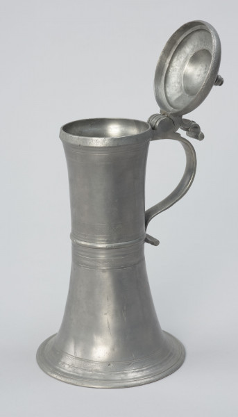 Cynowy dzban o kształcie cylindrycznym - Ujęcie z lewej strony, z ukazaniem części wlewowej; Wysmukły dzban o kształcie cylindrycznym z lekko wklęsłym brzuścem, z pierścieniowym zgrubieniem w centrum. Rozszerzająca się ku dołowi podstawa przechodzi w profilowaną stopę. Ucho taśmowe, silnie esowato wygięte, zakończone wałkiem. W górnej części ucha zawias podtrzymujący pokrywę z uchwytem wygiętym esowato. Pokrywa kopulasta, dwustopniowa z profilowaną, toczoną gałką do podnoszenia na szczycie. Na brzuścu u podstawy, pośrodku i przy górnej krawędzi, a także na pokrywie delikatne profilowane i ryte pasy.