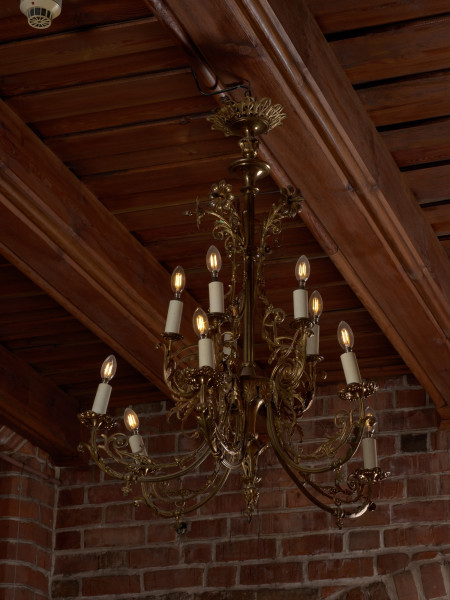 żyrandol - Ujęcie z lewej strony. Żyrandol 12-ramienny, mosiężny z kryształowymi soplami.Trzon w górnej części o przekroju kwadratowym, ozdobiony wypukłą dekoracją o formach geometrycznych i roślinnych, w dolnej części tralkowy z zakończeniem w kształcie szyszki. Ramiona w formie gałązek wygiętych półkoliście, połączonych przy trzonie w 4 wiązki po 3 elementy: dwa dłuższe po bokach i krótsze w środku. Każda trójka przechodzi przy trzonie w jedną spiralnie zwiniętą gałązkę akantu. Środkowe części ramion ozdobione są analogicznymi motywami: łodyżką z listkami rozszczepiającą się na dwa spiralnie skręcone pędy. Profitki ażurowe, zbudowane z ułożonych w rozetkę listków akandu. Przy górnej części trzonu, w połowie wysokości motyw dekoracyjny złożony z dwóch wygiętych, a na końcach spiralnie skręconych gałązek akantu. Górny talerz maskujący pod sufitem zawieszkę - płaski w formie kwiatu akantu. Ramiona i trzon połączone luźno zwieszonymi łańcuszkami, umieszczonymi także przy talerzu podsufitowym i ozdobach górnej części trzonu. Przy profitkach szklane wisiorki: 16 sztuk szlifowanych w kształcie sopelków - podłużne o trójkątnym przekroju z ostrosłupowym zakończeniem oraz 5 sztuk w kształcie pękatych łezek. Świece tulejowe z tworzywa sztucznego, zaopatrzone we wkręty do żarówek świecowych. Wewnętrzna instalacja elektryczna.