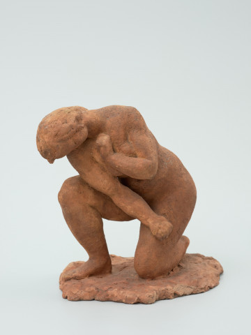 akt kobiecy - Ujęcie z przodu; Rzeźba postaci kobiecej z czerwonej terakoty. Kobieta klęczy na lewym kolanie, prawą rękę opiera na lewym udzie, a lewą zasłania pierś. Głowę przechyla w kierunku prawego ramienia. Całość w tonacji pomarańczowo-czerwonawej.