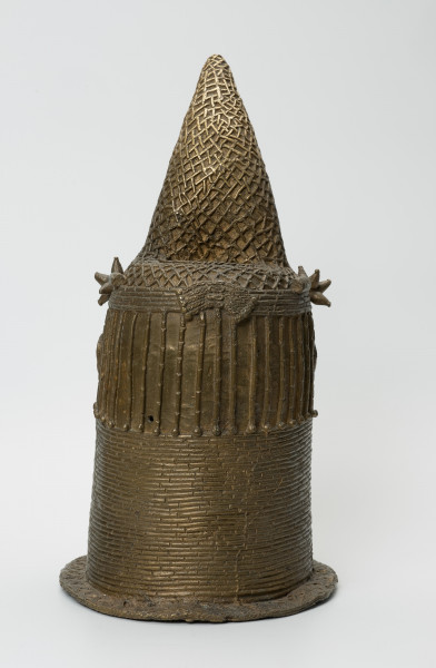 Głowa Królowej Matki - Ujęcie z tyłu. Brązowy, pusty w środku odlew głowy kobiety, o złotym zabarwieniu. Twarz o cechach negroidalnych, wydatnych, lekko rozchylonych ustach oraz szerokim u nasady nosie ma oczy z wyraźnie zaznaczoną linią powiek. Na ich wysokości usytuowane są nieduże uszy. Lekko wypukłe czoło dekorowane jest pionowymi inkrustacjami z żelaza oraz trzema podłużnymi, symetrycznie rozmieszczonymi, pogrubieniami nad każdym okiem. Szyję i podbródek do wysokości dolnej wargi zasłonięto masywnym naszyjnikiem z korali. Głowa jest nienaturalnie poszerzona w tylnej części, tuż pod przechyloną do przodu koroną o charakterystycznym kształcie dziobu papugi, którą nad uszami ozdobiono dwoma rozetami. W dolnej części korony widnieje pięć ozdób o walcowatej formie, trzy nad czołem, dwie za rozetami oraz opaska z korali. Tuż za koroną znajduje się duży, owalny otwór na rzeźbiony kieł słonia, którego brak. Poniżej, po obu stronach twarzy i z tyłu głowy, zaznaczono zaplecione w warkoczyki włosy. Z tyłu głowy pomiędzy włosami a naszyjnikiem usytuowany jest niewielki, okrągły otwór. U podstawy odlewu znajduje się pozioma listwa, dekorowana motywem plecionki.