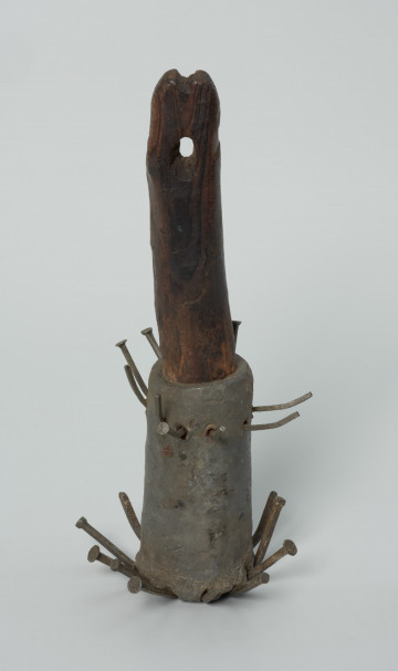 Kotewka - Ujęcie z przodu w pionie. Narzędzie pomocnicze niewielkich rozmiarów, wykonane techniką domową. Ołowiana tuleja osadzona na wykonanym z drewna, dość krótkim, prostym trzonku. W  górnej powierzchni trzonka płytkie nacięcie, poniżej niego elipsowaty otwór służący do przeciągania linki. W tulei mocowane dwa rzędy, odchodzących od niej promieniście, wygiętych ku górze metalowych gwoździ (łącznie 23 szt.). Niektóre z gwoździ pozbawione są główek.