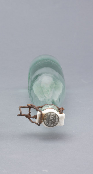 szklana butelka z kapslem - Ujęcie z góry; Butelka piwówka z grubego szkła przeźroczystego półbiałego z odcieniem zielonym. Szyjka przechodząca łagodnie z korpusu zakończona przegrubieniem z wklęską na drut zamknięcia bocznego - dźwigniowego. Kapsel z białego porcelitu owalny z wypustami na dwóch bokach.