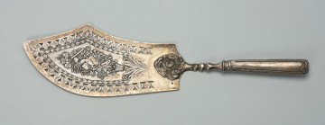 srebrna łopatka o kształcie zbliżonym do tasaka - Ujęcie z przodu; Srebrna łopatka - nóż do ryb o kształcie zbliżonym do tasaka (jedna strona obła, druga wycięta w kształcie nierównobocznego trójkąta) z centralnie umieszczonym trzonkiem. Wzdłuż brzegu powyżej ornamentu zdobiącego uchwyt trzonka pas ażurowego i grawerowanego ornamentu w formie listków, od wewnetrznej strony wzbogaconego rzędem drobnych, potrójnych, wypukłych listków. Pośrodku na delikatnie fakturowanym tle repusowana kompozycja złożona z akcesoriów i narzędzi ogrodniczych (grabie, konewka, łopata, kapelusz z kokardą) ułożonych wśród roślin (kłosy zbóż, gałązki winnej latorośli, gałązki z liśćmi i kwiatami) w wydłużony bukiet wstawiony do umieszczonego poniżej koszyka z kwiatami. Pod nim bukiet o wąskich, wydłużonych kwiatach z trzema symetrycznie rozłożonymi kwiatami. W dolnej części łopatki o ostro wyciętych narożnikach bogata, silnie grawerowana i repusowana dekoracja tworząca wypukły, owalny zwój liści, kwiatów i owoców, których skraj wtopiony jest w trzonek uchwytu. Dolna część uchwytu dolutowana rączka z półwałkowym cięciem w górnej części, głęboko trybowanym profilem po bokach i sercowym zakończeniem wewnętrznego profilowania od dołu.