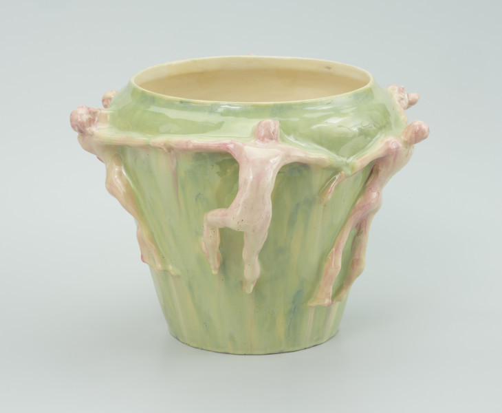 ceramika - Ujęcie z boku; Postacie ceramiczne przyczepione do malowanego wazonu. Widoczna postać męska zwrócona tyłem do nas ciągnąca po bokach postacie. Mężczyzna zapiera sie nogami i głową o wazon, z wypiętymi pośladkami. Postacie malowane kremowo z domieszką czerwonej barwy.