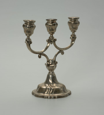 srebrny świecznik trójramienny - Ujęcie z przodu, z lewej strony; Świecznik srebrny trójramienny, neorokokowy. Stopa kielichowa o falistym narysie cokoliku i płaszczu zdobionym czterema potrójnymi wybrzuszeniami o falistych liniach, zbliżających się do siebie ku górze. Trzon w formie nodusa przykręconego do stopy. Nodus w dolnej części dekorowany analogicznym puklowaniem jak stopa, w górnej części gładki. Do nodusa dołączone dwa wsporniki w formie litery C (uproszczonego roccailla), na których górnych częściach spoczywają dwa ramiona w formie leżących liter C, wspierające krótkie trzony opraw do świec, dekorowane czterema podwójnymi puklowaniami o falistej linii. Wewnątrz opraw tulejki do świec z płaską profitką (w środkowej oprawie brak wewnętrznej tulejki). Środkowa oprawa z dodatkowym zakończeniem trzonu umieszczona została pomiędzy zgrubionymi końcówkami ramion.