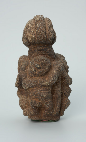 rzeźba; przedmiot obrzędowy; figura kultu zmarłych - Ujęcie z tyłu. Rzeźbiona w ziemistym steatycie siedząca postać ludzka o cechach kobiecych, otoczona przez trójkę mniejszych, dzieciecych postaci, skierowanych ku niej z uniesionymi rękoma.