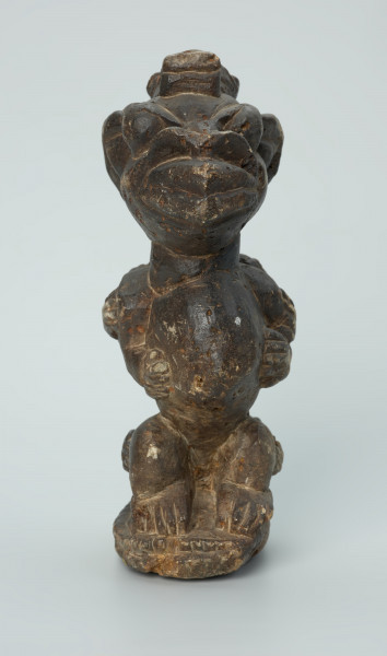 rzeźba; przedmiot obrzędowy; Figura kultu sił wegetacji - Ujęcie z przodu. Rzeźbiona w grafitowym steatycie, siedząca na owalnej podstawie, postać ludzka, którą od tyłu obejmuje postać zoomorficzna o cechach jaszczurki.