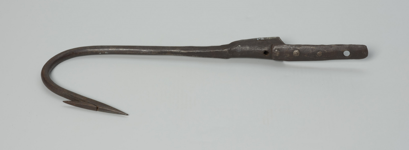 harpun - Ujęcie z przodu w poziomie. Duży, wykonany z żelaza techniką kowalską harpun. Część pracująca łukowato wygięta, zaostrzona z przygwintowanym pojedynczym, ruchomym  zadziorem. Zakończona tuleją, w której osadzona była rękojeść (rękojeści brak).  Z tulei wyprowadzony krótki pas metalowy, do którego za pomocą trzech nitów, po zewnętrznej stronie, mocowany jest pas dłuższy. W tulei i w dłuższym pasie wycięte, znajdujące się na jednej linii, okrągłe otwory.