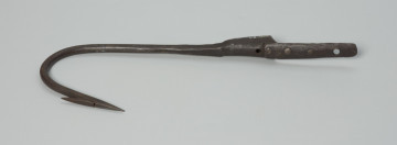 harpun - Ujęcie z przodu w poziomie. Duży, wykonany z żelaza techniką kowalską harpun. Część pracująca łukowato wygięta, zaostrzona z przygwintowanym pojedynczym, ruchomym  zadziorem. Zakończona tuleją, w której osadzona była rękojeść (rękojeści brak).  Z tulei wyprowadzony krótki pas metalowy, do którego za pomocą trzech nitów, po zewnętrznej stronie, mocowany jest pas dłuższy. W tulei i w dłuższym pasie wycięte, znajdujące się na jednej linii, okrągłe otwory.