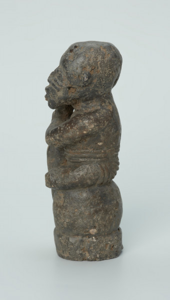 figura kultu przodków - Ujęcie z boku z lewej. Rzeźbiona w grafitowym kamieniu postać ludzka w pozycji siedzącej - najprawdopodobniej mężczyzny. Postać siedzi na wysokiej podstawie, która swym kształtem przypomina kamień. Rzeźba ma charakterystyczne poziome nacięcia widoczne na obu policzkach. Widoczne rysy, mikropęknięcia i liczne malutkie otworki.