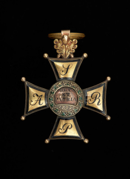 Order Virtuti Militari III klasy - Ujęcie rewersu bez widocznego uszka. Na rewersie ramion krzyża inicjały. W środkowej, okrągłej tarczy napis w 4 wierszach. Wokół wieniec laurowy z zielono emaliowanymi liśćmi. U góry gałązka dębu z uszkiem, przez które przechodzi kółko.