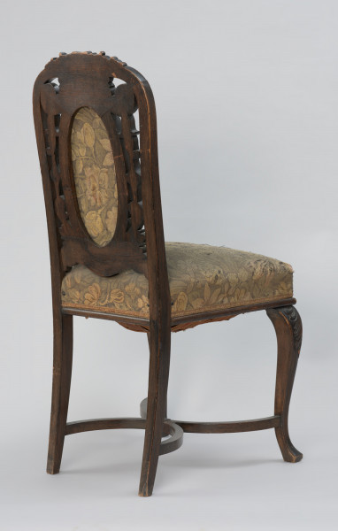 krzesło - Ujęcie z tyłu, z lewej strony. Krzesło w stylu neobarokowym, oparcie i siedzisko tapicerowane. Nogi zwężające się do dołu, przednie wygięte kabriolowo, górne wybrzuszenie ozdobione płaskorzeźbionym motywem palmety, zakończenie dolne trójkątne. Tylne nogi lekko wygięte, powiązane są z przednimi krzyżową poprzeczką umieszczoną u dołu, składającą się z dwóch stykających się w środku półokrągłych pałąkowych listew. Konstrukcja zaplecka stanowi pionowe przedłużenie tylnych nóg o profilowanych krawędziach, zamykających się dwoma półkołami w kształt serca, ponad którym wyprowadzono łukowe zwieńczenie w formie mięsiście rzeźbionego, rozłożonego na boki bukietu liści. Poniżej sercowego styku listew bocznych wydłużony w pionie medalion opasany płaską listwą z wypełnieniem tapicerką. Tkanina w kwiatowy wzór w odcieniach zieleni, różu, żółcieni i ciemnego popielu. Pomiędzy owalem medalionu i listwami oparcia ażurowe wypełnienie mięsistą płaskorzeźbą o motywach roślinnych. Taką samą tkaniną obite jest siedzisko o konstrukcji sprężynowej, stosunkowo wysokie, poszerzające się do przodu, w dolnej części wykończone półwałkiem i pasmanterią, kryjącą krawędź tapicerki.