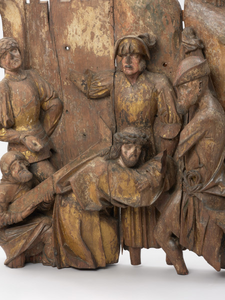 sztuka sakralna, kwatera ołtarza, płaskorzeźba - detal w lewym dolnym rogu; Płaskorzeźba przedstawiająca Chrystusa pochylonego pod ciężarem krzyża oraz pomagającego mu na rozkaz żołnierzy Szymona Cyrenejczyka. Scena na tle pejzażu górskiego przed murami miasta. Kwatera środkowa późnogotyckiego ołtarza pasyjnego z Wkryujścia (Ueckermünde).