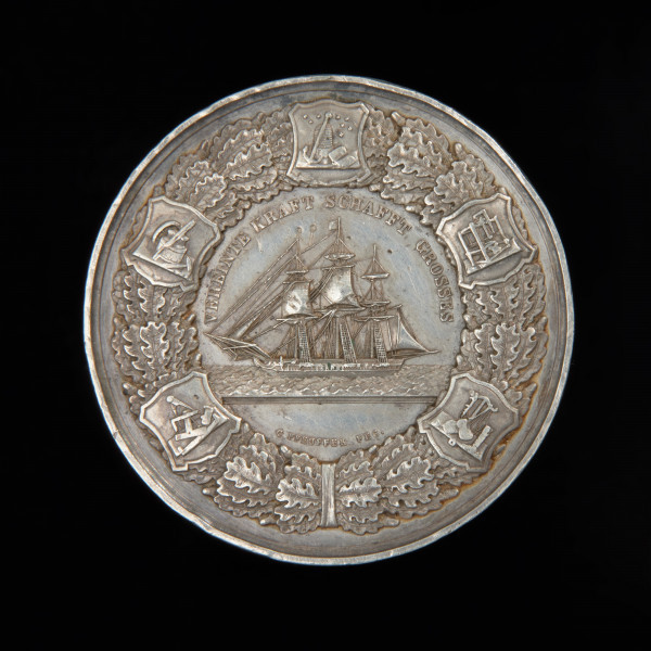 medal nagrodowy - Ujęcie awersu. Medal z wizerunkiem wspiętego gryfa na powalonym pniu i żaglowca na falach. Na rewersie trójmasztowy żaglowiec na falach w lewo, nad nim w łuku napis: zjednoczona siła stwarza wielkość, niżej w łuku sygnatura. W otoku wieniec dębowy z tarczami, w których symbole rzemiosła i przemysłu. Od lewej strony u dołu: stolarstwa, przemysłu maszynowego, pszczelarstwa i handlu, tkactwa oraz rymarstwa.