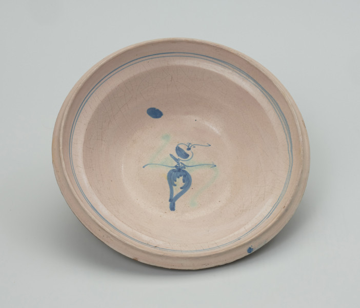 talerz - Ujęcie z góry z przodu. Talerz wykonany na kole garncarskim z gliny o jasnej barwie ( z małą zawartością tlenków żelaza). Dno niewyodrębnione przechodzi w lekko odchylony na zewnątrz korpus z wyraźnym kołnierzem. Szkliwiony tylko po wewnętrznej stronie i dekorowany odręcznie stylizowanym, jednoelementowym motywem roślinnym. W miejscu przejścia dna w korpus przypadkowa kropla farby. Wylew z kołnierzem zdobiony podwójną, miejscami niestaranna linią. Zdobienia w kolorach: dominującym niebieskim oraz zielonym.