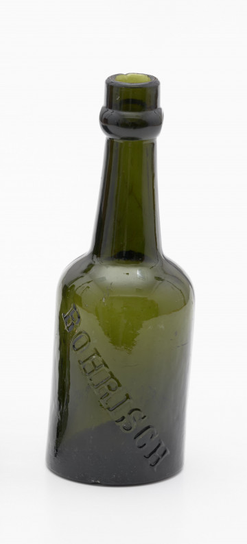 Butelka piwówka z grubego szkła o ciemnym kolorze oliwkowo-zielonym - Ujęcie z przodu; Butelka piwówka z grubego szkła butelkowego w ciemnym kolorze oliwkowo-zielonym. Szyjka wyodrębniona, lekko zwężająca się ku górze, poniżej wylewu pierścień z dwoma otworami na zamknięcie pałąkowe (brak). Dno wklęsłe.