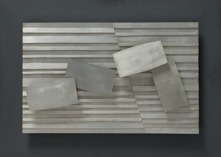 kompozycja abstrakcyjna - ujęcie z przodu; Kompozycja materiałowa o układzie prostokąta poziomego. Płyta metalowa łączona ukosem od lewej strony, w 1/3 szerokości. Na płaszczyźnie cztery elementy tego samego formatu (prostokąt o lekko wybrzuszonych dłuższych krawędziach) zestawione względem siebie pod równym kontem.