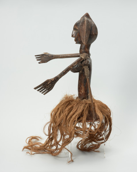 Ujęcie z lewego boku. Lalka umieszczona na koszyku-czapeczce pokrytej włóknami roślinnymi, noszona na głowie, przedstawiająca kobietę od pasa w górę. Linia brwi i nos wyodrębnione z twarzy. Usta lekko zaznaczone. Twarz ozdobiona skaryfikacją: po dwie linie pionowe na czole i policzkach. Ręce ruchome, poruszają się w linii poziomej za pomocą patyków umocowanych w 