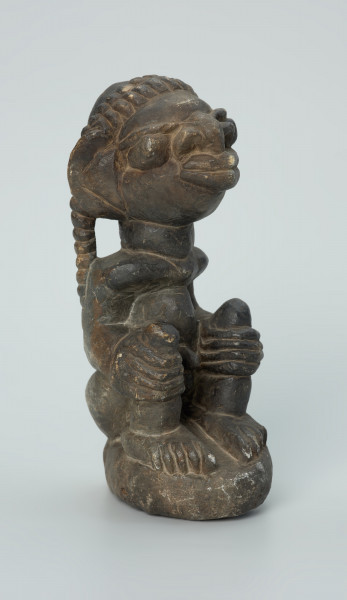 rzeźba; przedmiot obrzędowy; Figura kultu sił wegetacji - Ujęcie z przodu z prawej strony. Rzeźbiony w grafitowym steatycie, na owalnej podstawie, mężczyzna w pozycji siedzącej z opaską przedstawioną za pomocą wypukłego ornamentu kostkowego, której końce opadają na ramiona i plecy postaci.