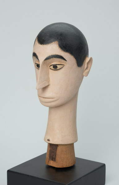 lalka teatralna: Tubabu Cè Kun - Ujęcie z przodu z lewej strony. Drewniane popiersie mężczyzny. Głowa owalna, zwężająca się ku dołowi. Zaznaczone krótkie włosy z falistą grzywką, pomalowane na czarno. Twarz i szyja pomalowane na blado-różowy, cielisty kolor. Zaznaczone czarne, łukowate brwi, migdałowe, wyrzeźbione oczy z powiekami i czarnym cienkim obramowaniem, z czarną okrągłą źrenicą w środku. Nos wąski, długi, usta lekko wypukłe, pomalowane na kolor cielisty. Szyja wąska, długa, rozszerzająca się ku dołowi.